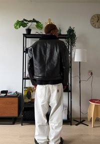 Blouson aviateur en cuir noir avec un col en fourrure, porté sur un pull noir. Associé à un pantalon blanc ample et des chaussures noires.