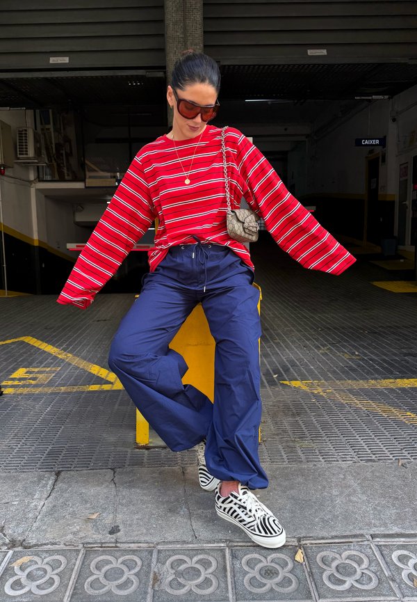 Mujer con camiseta de rayas rojas, pantalones azules y zapatillas de rayas sentada en una barrera amarilla en un aparcamiento, lleva gafas de sol y un pequeño bolso de hombro.