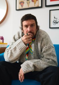 Homme aux cheveux courts et foncés et barbe, portant un pull gris et une écharpe colorée, assis sur un canapé bleu, le menton appuyé sur une main ornée de bagues.