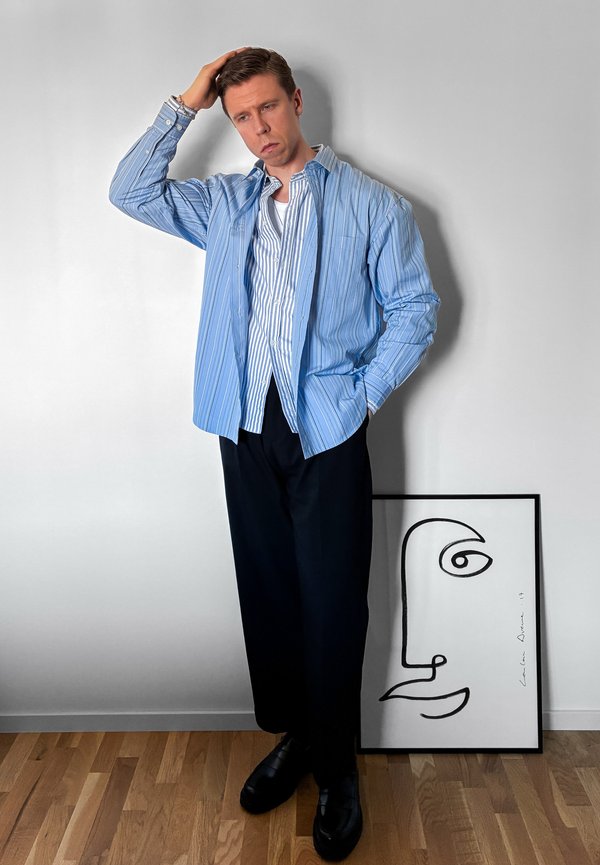 Tânăr în cămăși albastre cu dungi suprapuse și pantaloni negri stă cu mâna pe cap lângă un poster minimalist cu desen al liniei feței, sprijinit de un perete alb.