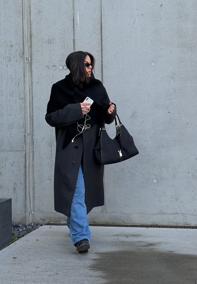 Abrigo negro de gran tamaño con capucha, jeans azules y bolso negro. Persona sosteniendo un teléfono inteligente con auriculares, de pie frente a una pared de hormigón.