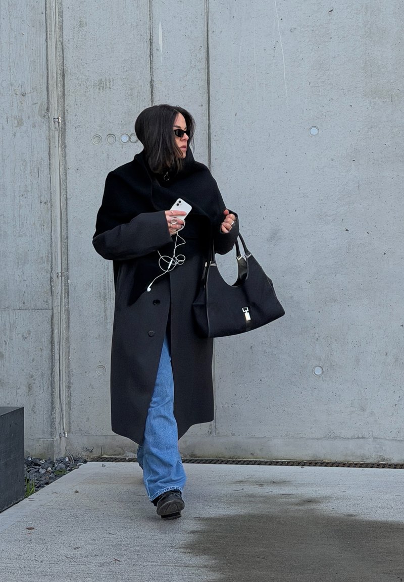Schwarzer übergroßer Mantel mit Kapuze, blaue Jeans und schwarze Tasche. Person hält ein Smartphone mit Ohrhörern und steht draußen vor einer Betonwand.