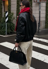 Melna ādas bumbvedēja jaka, sarkans šalle, krēmkrāsas kordurojas bikses un melna rokassoma. Stāv uz pārejas ar zaļojošu fona.