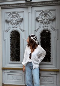 Camisa de linho branca com as mangas arregaçadas, jeans de cintura alta azul claro e um lenço padrão preto e branco, em pé ao lado de portas cinzentas ornamentadas.