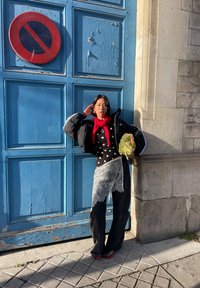 Γυναίκα που φοράει μαύρο μπουφάν φουσκωτό, κόκκινο κασκόλ, μαύρο τοπ με πουά και φαρδιά παντελόνια με δαντελένια λεπτομέρεια, στέκεται δίπλα σε μπλε πόρτα με πινακίδα απαγόρευσης στάθμευσης.