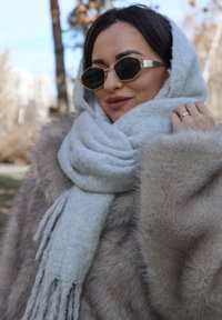 Szürke gyapjúsál egy bolyhos bézs szőrmekabát felett; kerek fekete napszemüveg arany díszítéssel; puha textúra és rojtos sálvégek.