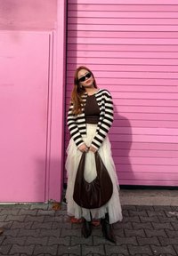 Cardigan à rayures noires et blanches porté sur un haut marron, jupe en tulle blanche superposée, sac hobo marron et bottes marron hautes jusqu'au genou devant un mur rose.