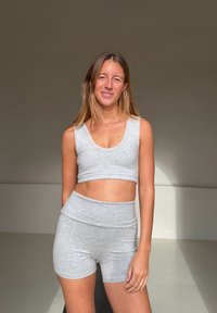 Jeune femme aux longs cheveux portant un crop top gris et un short, debout avec un sourire détendu sur un fond neutre uni.
