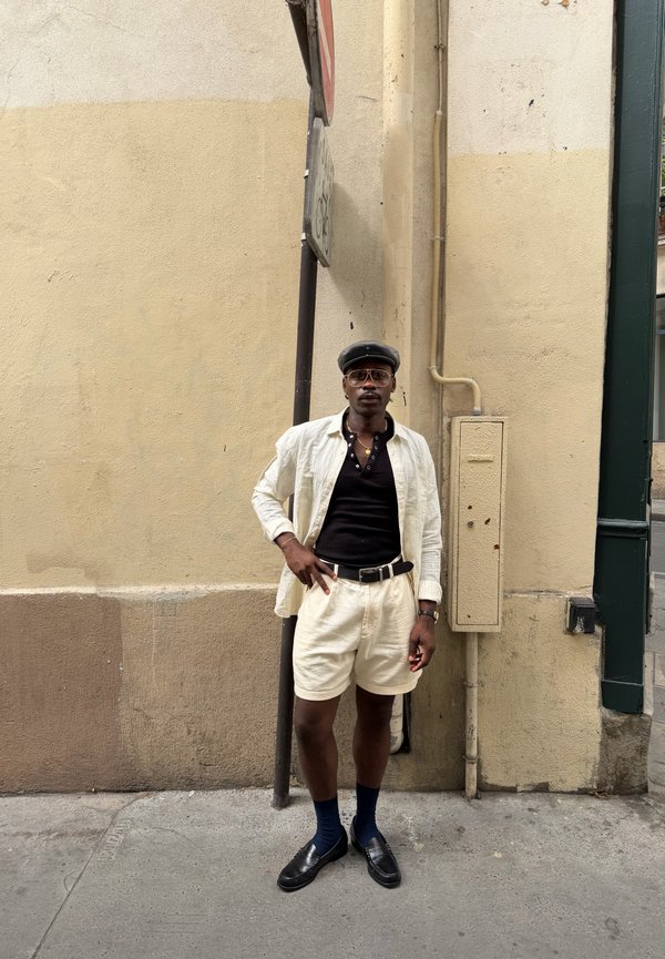 Homme portant une casquette noire, des lunettes, une chemise noire, une veste blanche ouverte, un short blanc, des chaussettes foncées et des chaussures noires, debout contre un mur beige dans la rue.