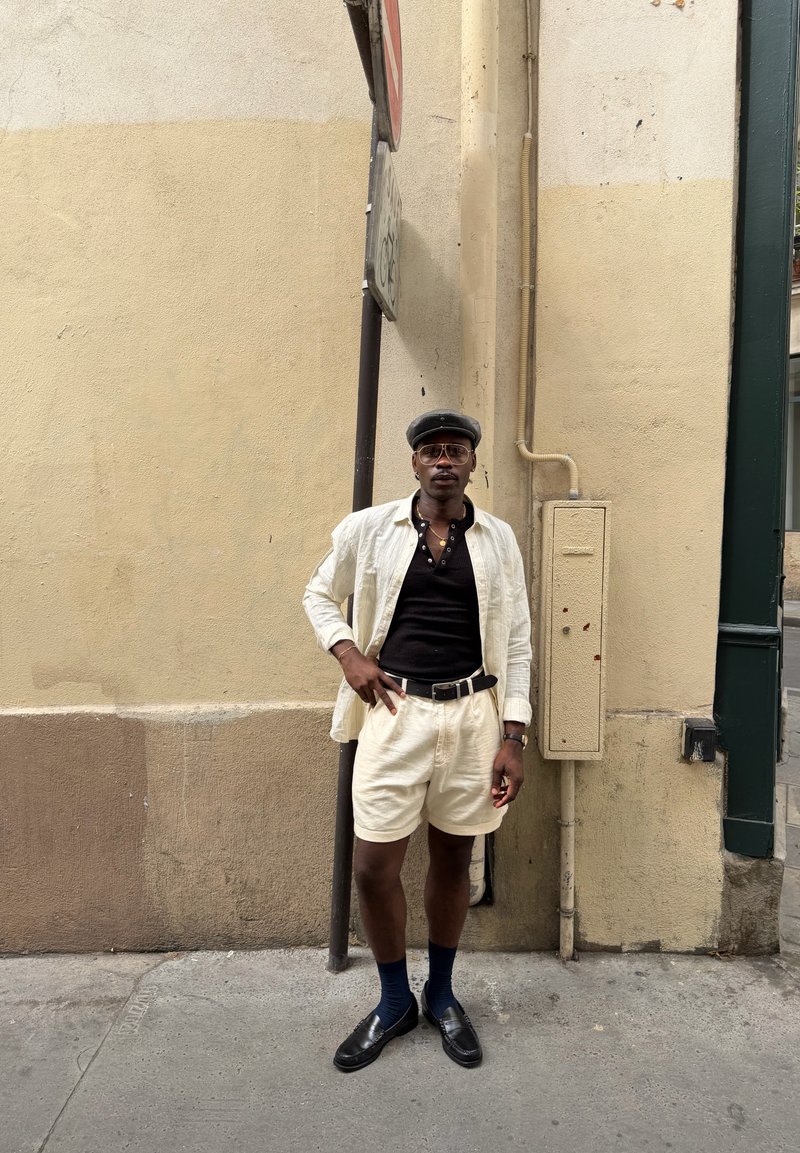 Homme portant une casquette noire, des lunettes, une chemise noire, une veste blanche ouverte, un short blanc, des chaussettes foncées et des chaussures noires, debout contre un mur beige dans la rue.