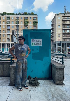 Camiseta gráfica sin mangas gris, vaqueros de mezclilla azules con un aspecto desgastado, zapatillas en blanco y negro, bolsa verde en la acera, ambiente urbano.