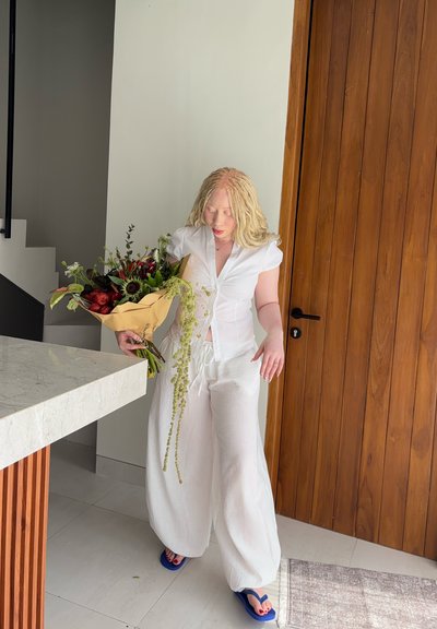 Persona con ropa blanca sosteniendo un gran ramo de flores mixtas cerca de una puerta de madera en un interior moderno y minimalista.