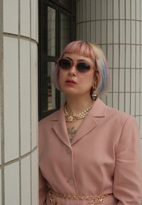 Person med kort pastel regnbuefarget hår, rosa solbriller, septumpiercing, perlekjede, hjerteørering, rosa blazer og gullkjede belte, stående ved en flislagt vegg.
