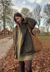 Tmavě zelená lehká bunda se zipem vpředu, dvěma bočními kapsami a hnědou koženou kabelkou přes rameno. Dlouhé svetrové šaty z měkké látky, tmavé punčochy.