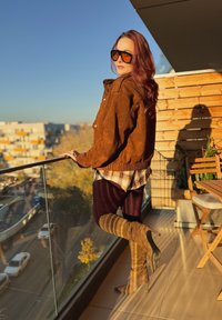 Giacca in suede marrone dal taglio comodo, camicia a quadri sotto, leggings color bordeaux e stivali alti tanfino alla coscia, in piedi su un balcone con ringhiera in vetro.