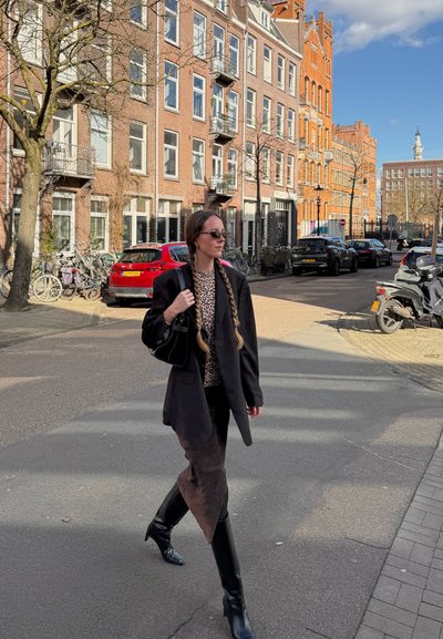 Mujer con trenzas y gafas de sol camina por una calle soleada en una zona urbana con edificios de ladrillo, coches estacionados, bicicletas y scooters.