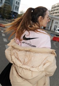 Giacca a vento beige sopra una maglietta Nike rosa chiaro con logo e testo neri. Capelli lunghi raccolti in una coda di cavallo, indossando occhiali.