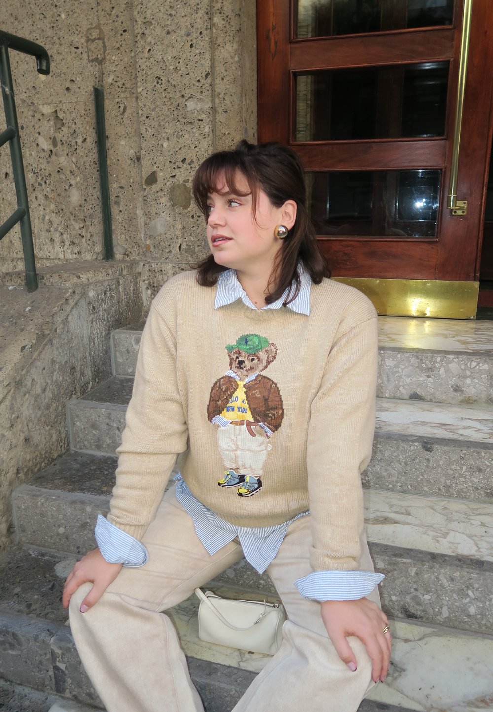 Beżowy sweter dzianinowy z motywem misia; niebieska koszula w paski pod spodem; kremowe spodnie; mała biała torebka; teksturowane kamienne schody.