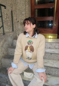 Beżowy sweter dzianinowy z motywem misia; niebieska koszula w paski pod spodem; kremowe spodnie; mała biała torebka; teksturowane kamienne schody.