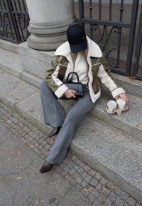 Pessoa com um casaco de inverno, calças cinzas e um gorro preto sentada em degraus de pedra, segurando uma mala preta e estendendo a mão para dois copos de café para levar.