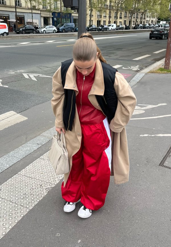 Jeune personne aux cheveux bruns attachés, portant un manteau beige, un gilet noir, un pantalon rouge et des baskets blanches, se tient sur un trottoir en ville tenant un sac beige.