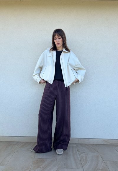 Chaqueta corta blanca con mangas grandes, camiseta negra debajo y pantalones anchos de rayas burdeos. Zapatos grises, de pie sobre un suelo de baldosas.