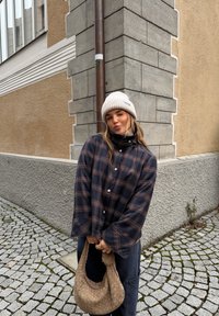 Chemise surdimensionnée à carreaux bleu marine et marron avec manches larges, associée à un bonnet en tricot beige et un sac à bandoulière tissé beige, sur une rue pavée.