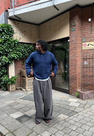 Jersey texturizado azul marino con mangas amplias, superpuesto sobre una camisa blanca, combinado con pantalones grises holgados y sandalias rojas, frente a una pared de ladrillos.