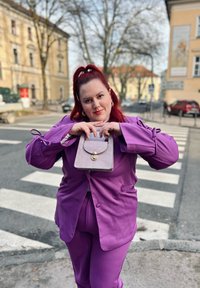Purple suit with a tailored fit, complemented by accessories. Carrying a light grey, structured handbag with a curved flap and round clasp.