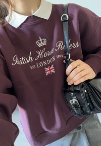 Granatna pulover s belim ovratnikom, na njem je napis "British Horse Riders", grafika krone in značka z Union Jack zastavo. Črna torbica z zaponko.