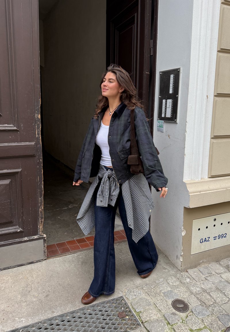 Casaco de xadrez preto e verde sobre uma blusa branca, jeans escuros de cintura larga e sapatos castanhos. Camisa xadrez atada na cintura.