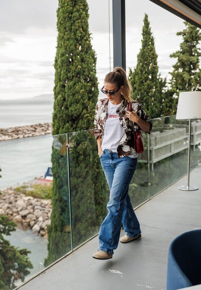 Chaqueta con un estampado floral sobre una camiseta blanca, jeans azules y zuecos beiges. Entorno exterior con barandilla de cristal y vegetación.