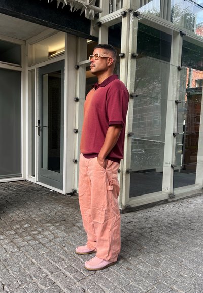 Una persona vestida con una camiseta polo burdeos y de gradiente naranja, pantalones cargo rosa claro y zapatos de lona rosa se encuentra de pie frente a un edificio de cristal.