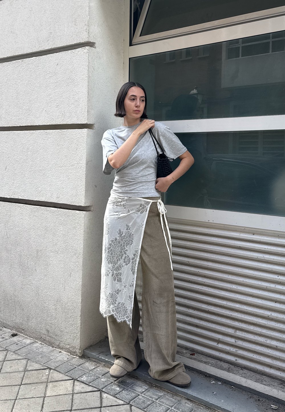 Camiseta gris, pantalones de pana anchos beige y una falda envolvente blanca de encaje. Bolsa de mano negra texturizada. De pie frente a una ventana moderna de cristal.