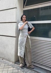 Camiseta gris, pantalones de pana anchos beige y una falda envolvente blanca de encaje. Bolsa de mano negra texturizada. De pie frente a una ventana moderna de cristal.