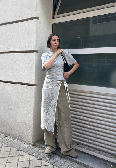 Camiseta gris, pantalones de pana anchos beige y una falda envolvente blanca de encaje. Bolsa de mano negra texturizada. De pie frente a una ventana moderna de cristal.