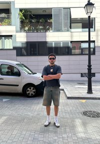 Vyras su akinių nuo saulės, juoda marškinėliais, džinsiniais šortais ir sportiniais bateliais stovi su sukryžiuotomis rankomis ant asfaltuotos gatvės šalia baltos mašinos ir lempinės stulpo.