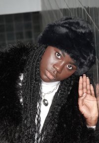 Mannequin portant un chapeau en fourrure noire et un manteau en fourrure noire, avec de longs cheveux tressés. Un collier chaîne en argent avec une pierre précieuse verte est visible.