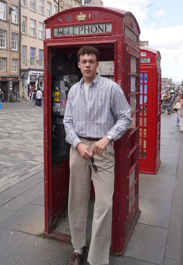 Jeune homme en pantalon beige et chemise rayée tient des lunettes de soleil à l'intérieur d'une cabine téléphonique britannique rouge dans une rue de la ville.