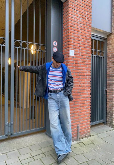 Persona vestida con una chaqueta de cuero negra sobre una sudadera azul, una camiseta a rayas y unos pantalones vaqueros anchos azules, apoyándose contra una pared de ladrillo con puertas de metal.
