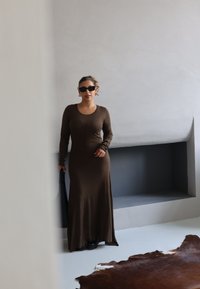 Mujer con un vestido maxi de manga larga marrón, usando gafas de sol negras, sosteniendo un bolso negro, de pie en una habitación minimalista con una alfombra oscura en el suelo.