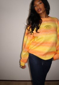 Pull ras du cou à motif dégradé orange et jaune avec un logo circulaire sur la poitrine. Associé à un jean bleu foncé.