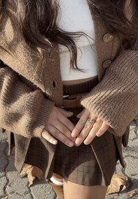 Cardigan marron tricoté à manches larges, haut blanc côtelé et jupe à carreaux marron. Accessoires comprennent une ceinture et une bague décorative.