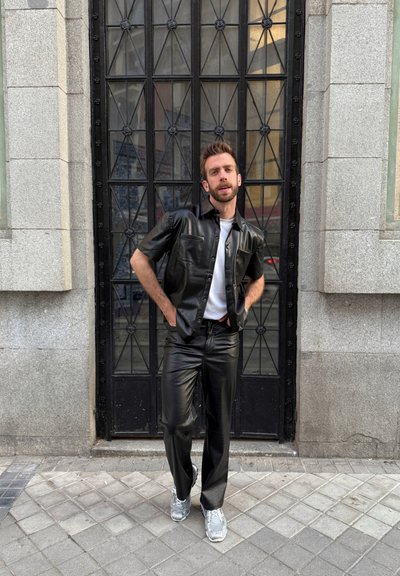 Camisa y pantalones de cuero sintético negros con una camiseta blanca debajo. Zapatillas plateadas. De pie frente a una puerta negra texturizada con un patrón en cuadrícula.