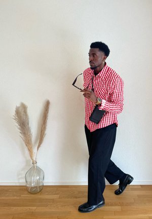 Hombre con una camisa de rayas rojas y blancas y pantalones negros sosteniendo gafas de sol, caminando en interiores junto a una pared blanca con un jarrón de cristal que contiene hierba de la pampas.