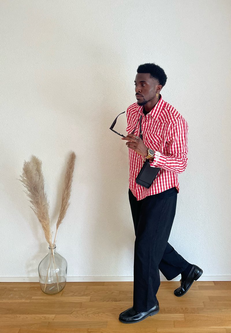 Homme en chemise rayée rouge et blanche et pantalon noir tenant des lunettes de soleil, marchant à l'intérieur près d'un mur blanc avec un vase en verre contenant des herbes de la pampa.