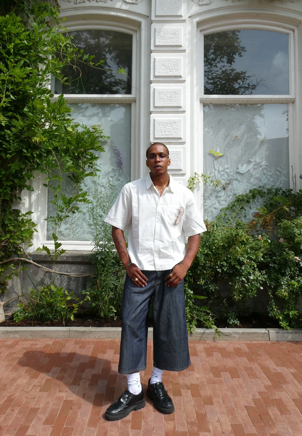 Hombre vestido con una camisa blanca de manga corta, pantalones de mezclilla anchos, zapatos negros, de pie sobre un pavimento de ladrillos frente a un edificio blanco con grandes ventanas y vegetación.
