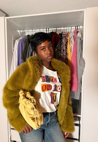 Νέος με κοντά μαύρα μαλλιά, που φοράει κίτρινο πανωφόρι με συνθετική γούνα, λευκό μπλουζάκι με σχέδιο, μπλε τζιν και κρατά μια κίτρινη τσάντα με πτυχώσεις.