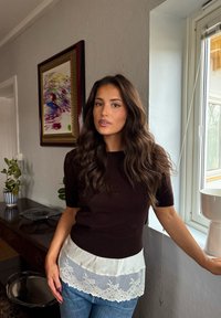 Joven con cabello largo y ondulado que lleva una blusa oscura con borde de encaje, de pie junto a una ventana en una habitación con una pintura abstracta enmarcada y plantas.