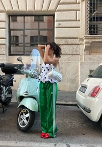 Λευκή μπλούζα με ώμους, με μαύρες βούλες, πράσινα παντελόνια φόρμας με λευκές ρίγες, κόκκινα ψηλοτάκουνα και μια μικρή λευκή τσάντα, δίπλα σε μια μέντα σκούτερ.